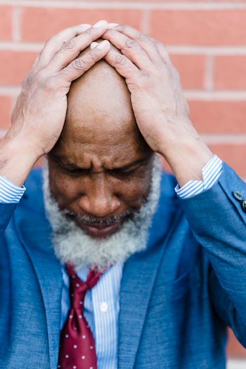 Free Worried bearded African American male entrepreneur in formal clothes standing near brick wall with hands on head after hard workday Stock Photo