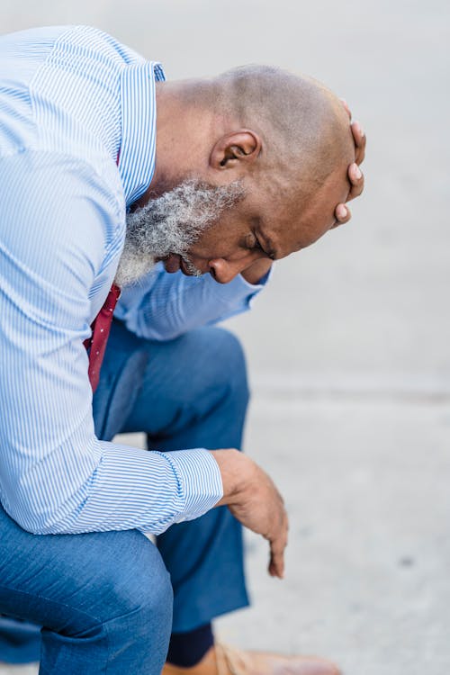 Side view of unhappy African American male entrepreneur in formal attire sitting on street with hand on head after workday
