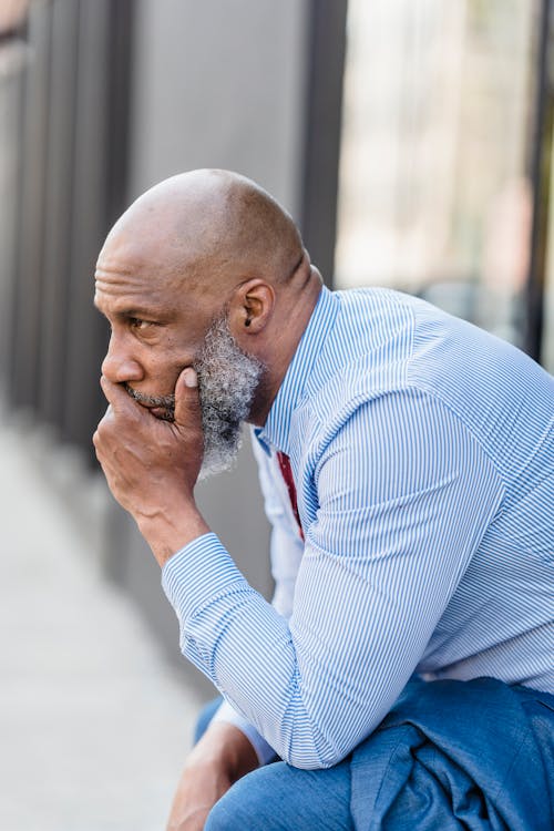 Side view of frustrated bearded African American businessman in formal wear sitting on street near building in city after workday