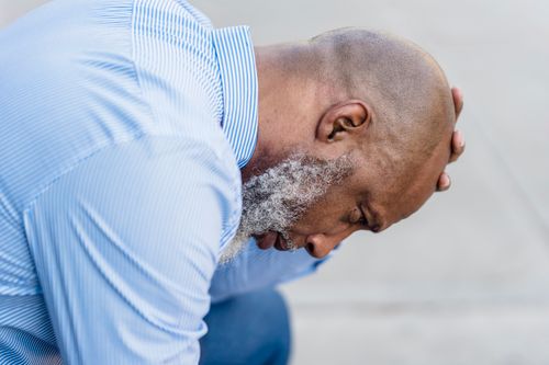 Free Side view of upset African American male worker with gray beard sitting on street with hand on head after workday Stock Photo