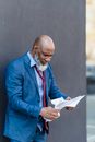 Confused African American male entrepreneur in formal suit reading applications in hands while standing near wall on street in city