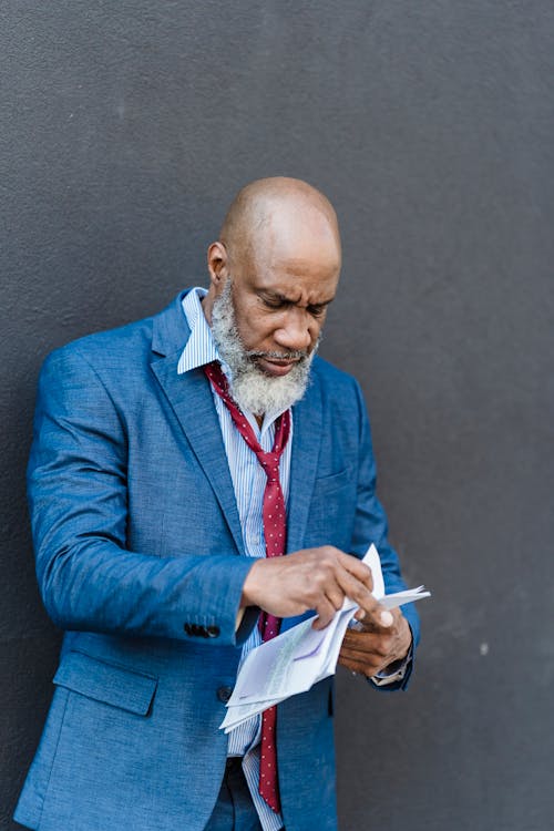 Upset bearded African American businessman in classy suit with official papers in hands standing on gray background on street of city
