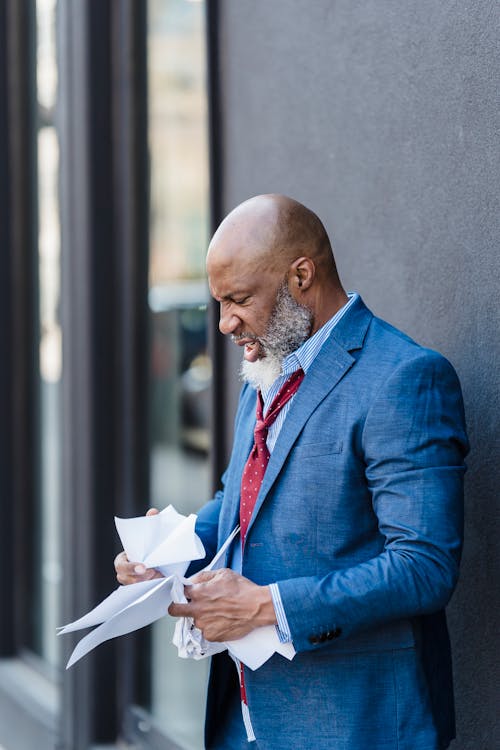 Free Side view aggressive African American male entrepreneur reading documents and crumpling paper while standing near building on street in city Stock Photo