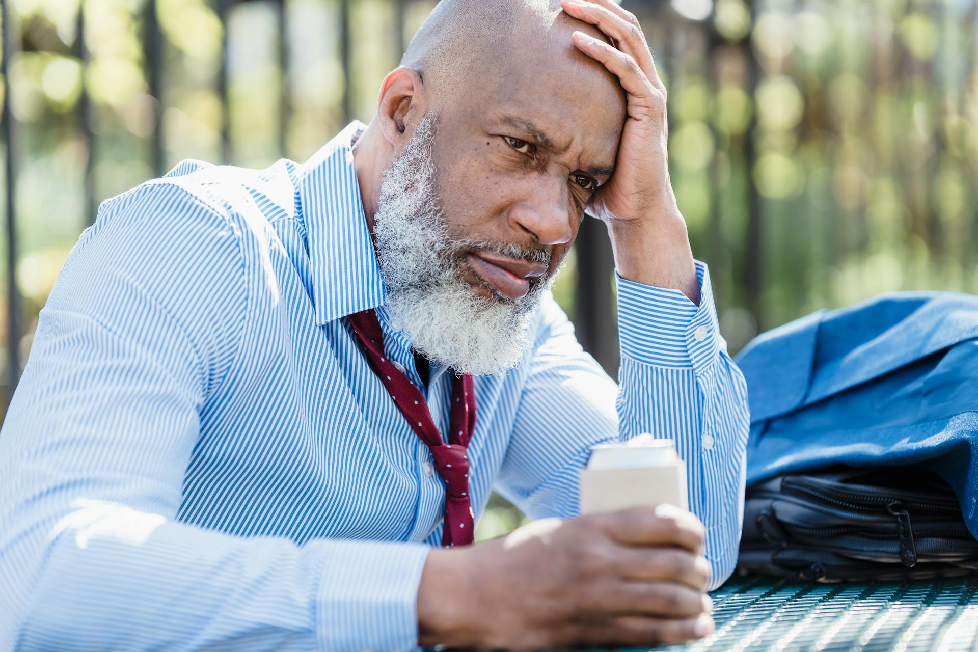 An adult man in business attire appears stressed while sitting outdoors with a drink.