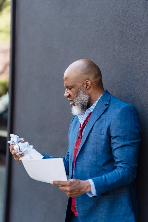 Free Side view of discontent African American businessman in formal wear making crumple of document while standing near wall on street Stock Photo