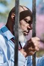 Serious concerned African American male in classy outfit gripping metal fence bars in sunlight