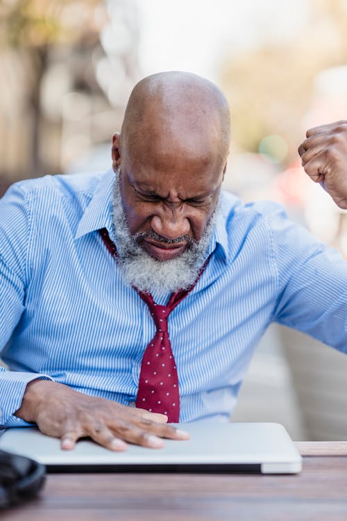 Elderly bearded African American male entrepreneur clenching hand into fist and closing laptop sitting at wooden table