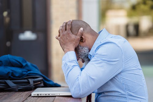 Side view of pensive middle aged African American male entrepreneur wearing blue shirt grabbing head with hands while sitting at table with closed laptop in veranda