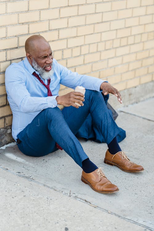 Full body of troubled senior bearded African American male in trendy outfit sitting on concrete pavement at brick wall with drink in tin can