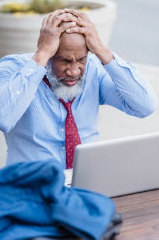 Stressed elderly black man grabbing head sitting with laptop