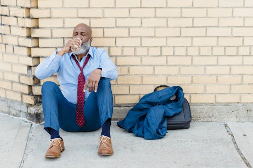 Full body of mature African American bearded businessman in blue trousers and light shirt with maroon tie sitting on ground at brick wall and drinking beverage from tin can