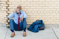 Full body of mature African American bearded businessman in blue trousers and light shirt with maroon tie sitting on ground at brick wall and drinking beverage from tin can