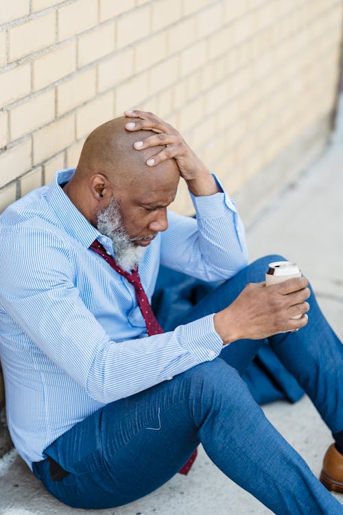 Senior troubled African American bearded businessman in shirt with tie and trousers sitting on ground near brick wall with bent knees and tin can of drink in hand
