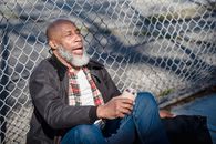 African American senior man with closed eyes and opened mouth sitting near metal fence and holding beer can
