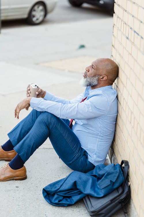 Free Side view of black man in formal clothes holding beer can while sitting on asphalt and leaning on brick wall Stock Photo