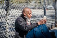 Side view of black homeless bald man sitting on street with beer can in hand and leaning on wire railing