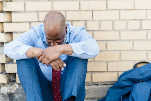 Sad shaved African American man in t shirt sitting with joined hands on background of brick wall and crying