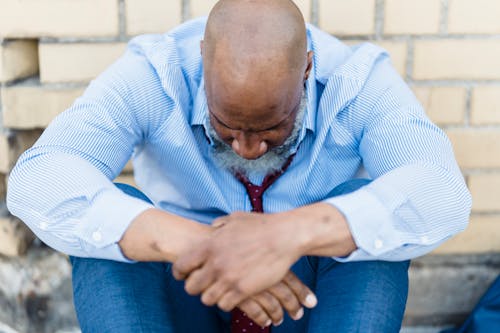 Hairless senior ethnic male in trendy outfit sitting near street wall and looking down in crisis