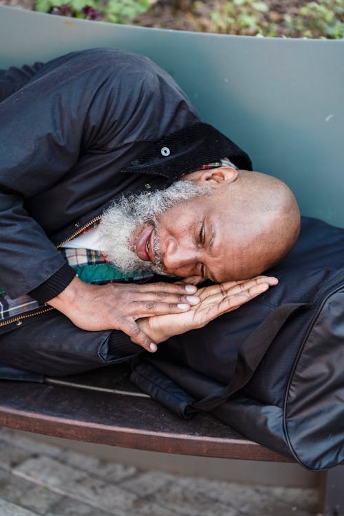 From above of senior homeless black male with white beard sleeping on bench in park