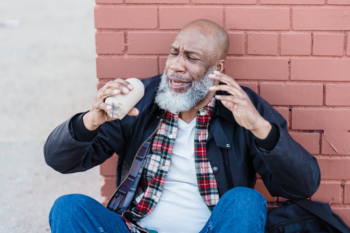 African American bearded male in casual outfit raising arms with beer can while resting on street near brick wall