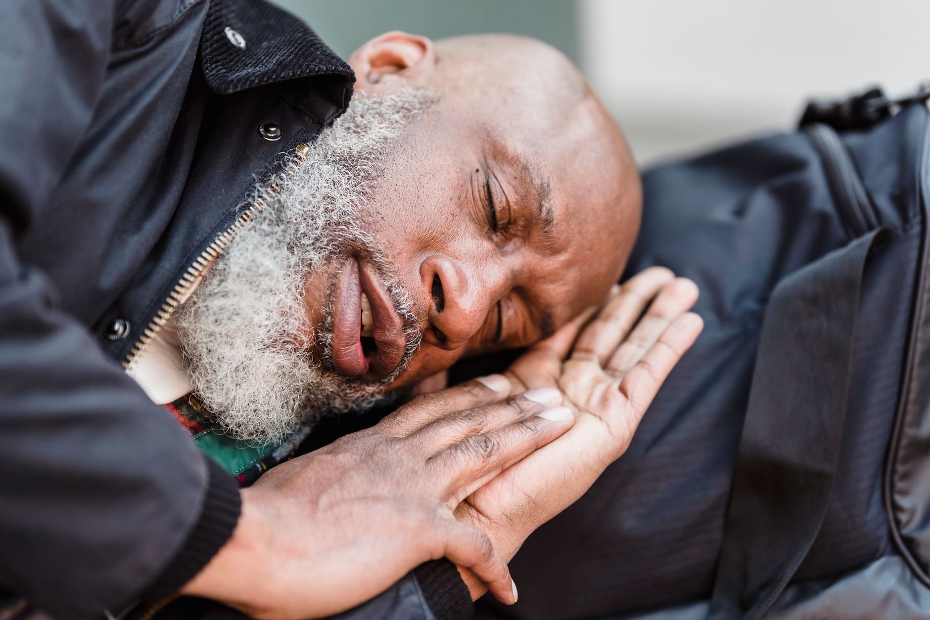 Ultra Bann A bearded elderly man sleeps comfortably outdoors with a backpack as a pillow.