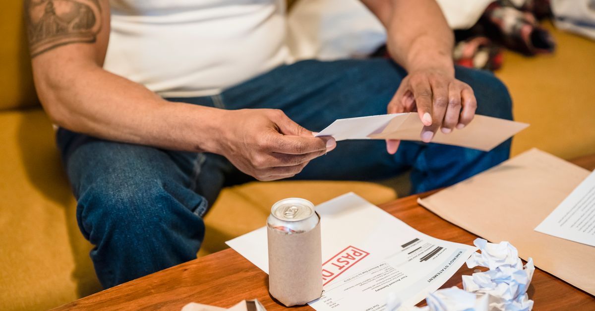 A man inspects his mail while seated on a couch, envelopes and papers scattered.