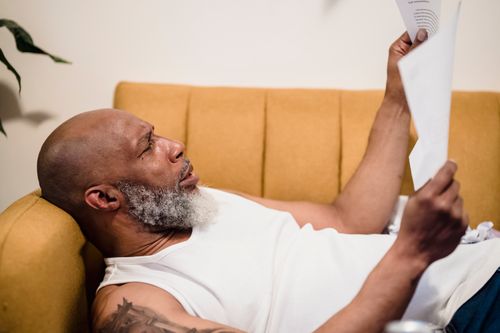 Free Bald man with facial hair lies on a mustard couch, reading documents in a relaxed setting. Stock Photo