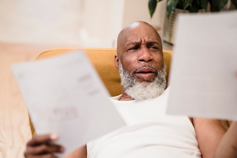 An older Black man with a white beard sits back in a yellow armchair at home, frowning with concern as he reads two paper bills, one in each hand.