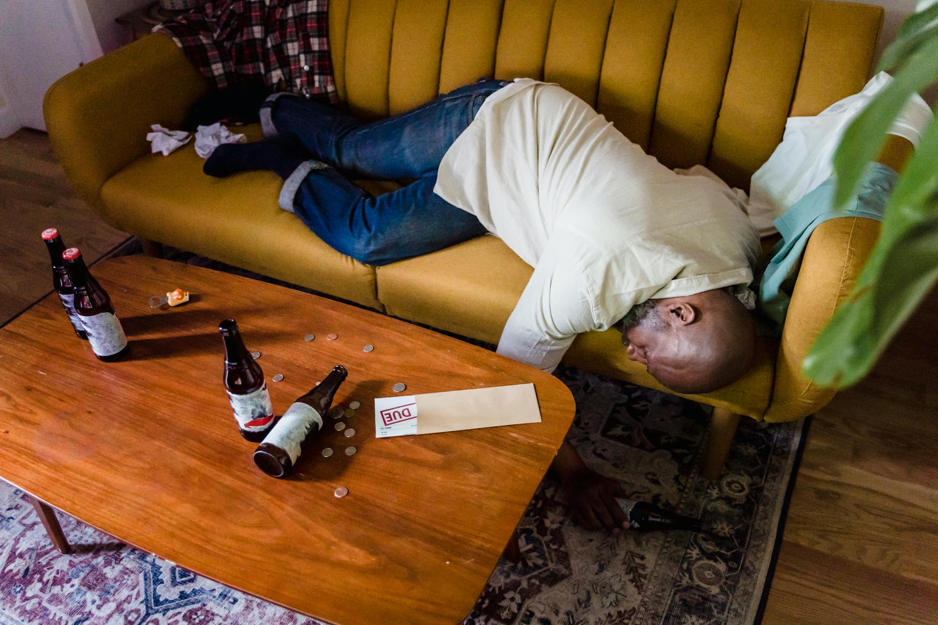 A man sleeping on a couch with empty bottles and an overdue bill, symbolizing financial stress and exhaustion.