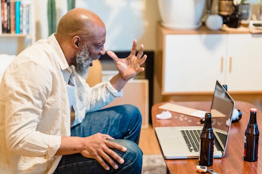 Bald man angrily gestures at laptop while sitting indoors with beer bottles around.