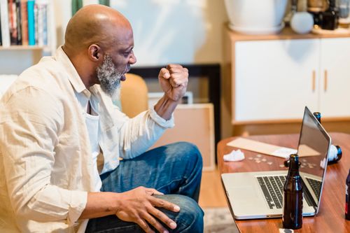 Free Adult man cheering during a video call, indoors with a beer bottle on wooden table. Stock Photo