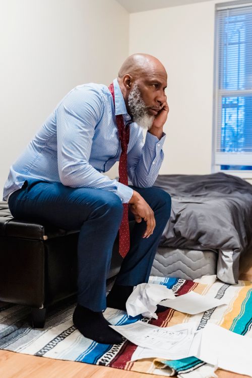 Free African American man sitting stressed with scattered papers in a home interior. Stock Photo