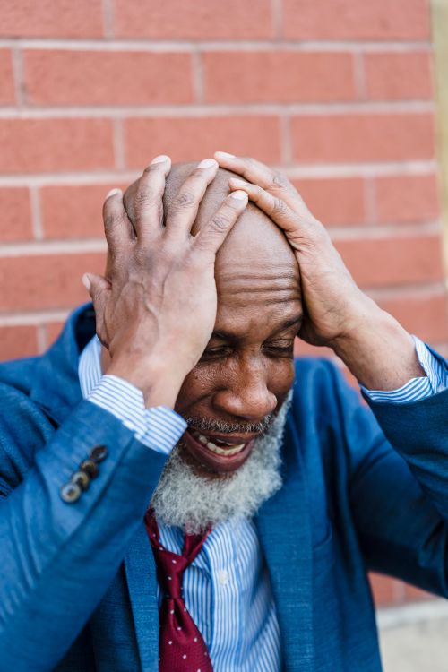 Free Senior man in a blue suit with hands on head, expressing stress outdoors against a brick wall. Stock Photo