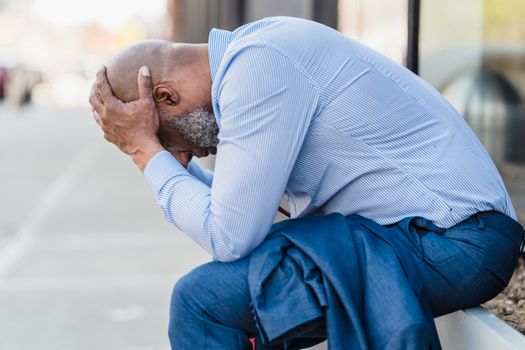 An adult man sitting outdoors, showing stress and contemplation with his head in hands.