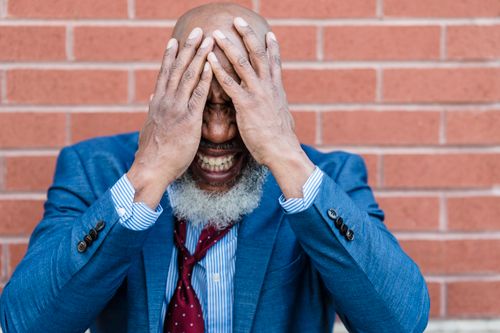 Free A bearded man in a blue suit looks frustrated, covering his face with his hands in front of a brick wall. Stock Photo
