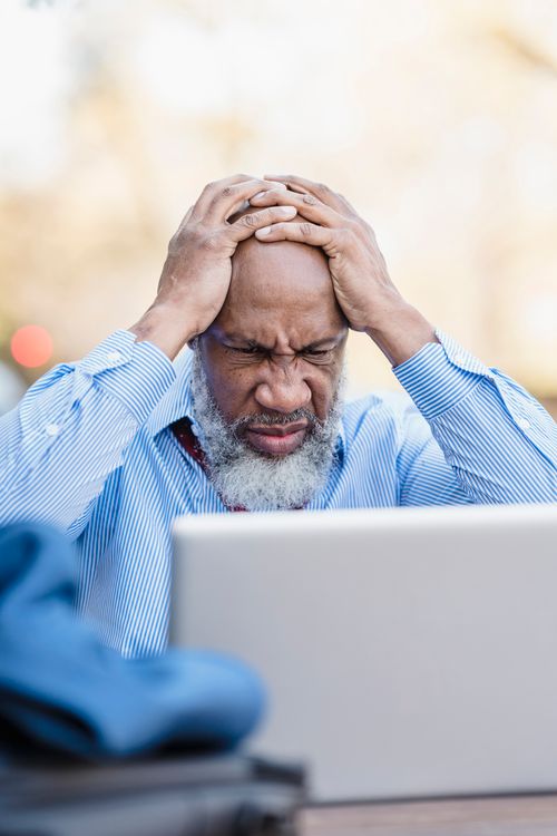Free Senior man sitting outdoors with head in hands, appearing stressed while using a laptop. Stock Photo