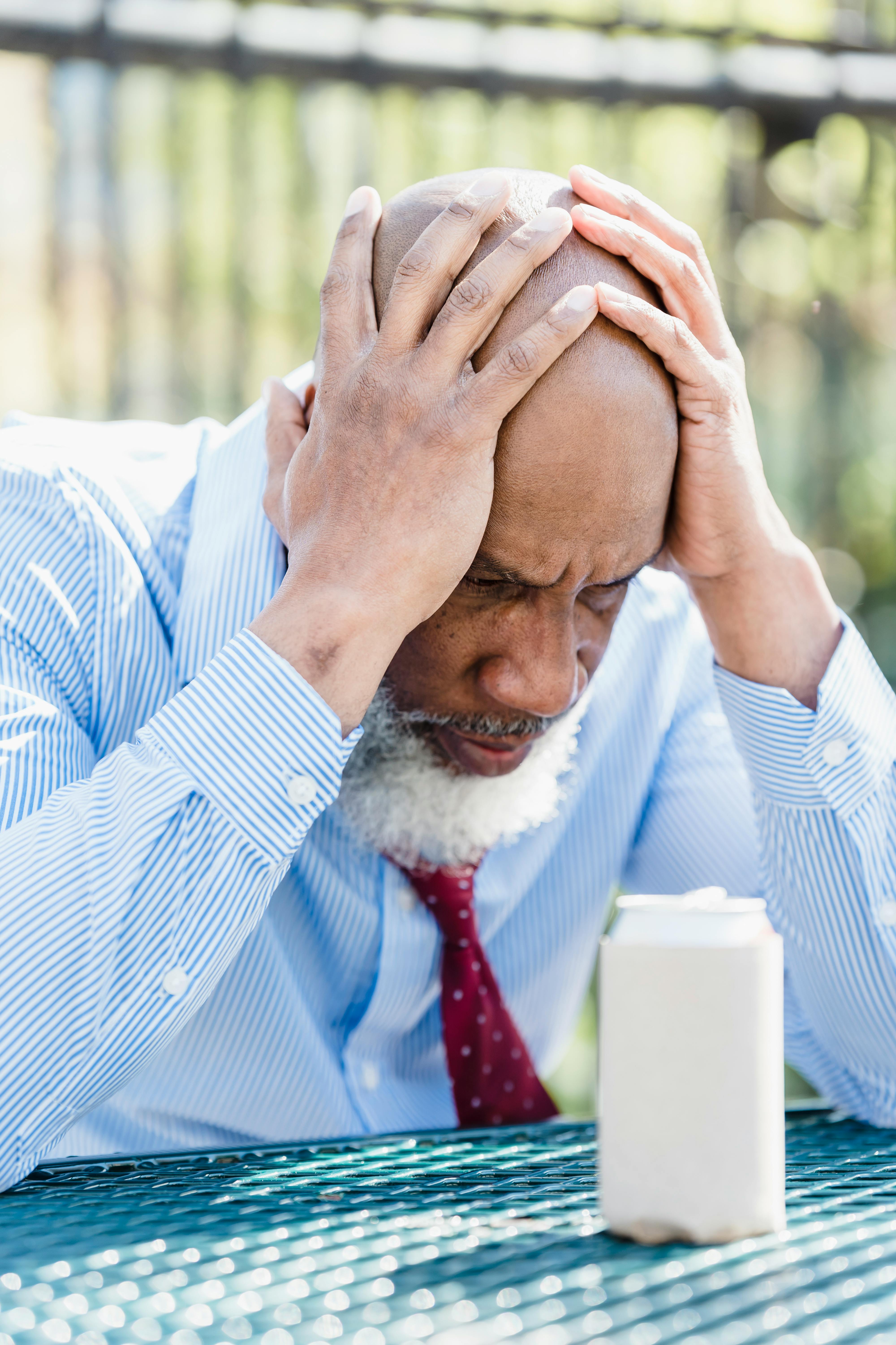 A bearded man looks pensive while sitting outdoors, holding his head.