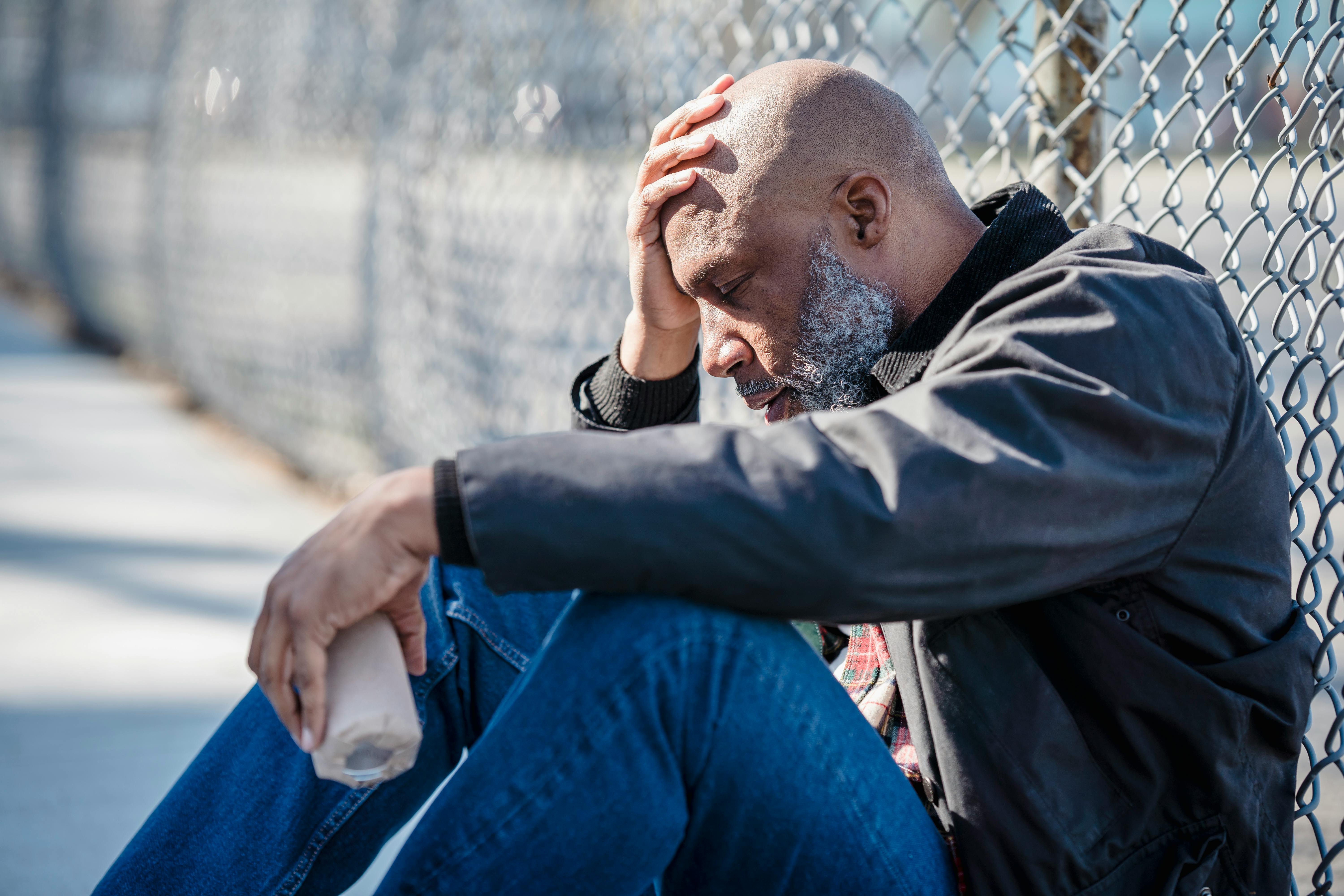 Pensive adult man with hand on head sitting by a fence, holding bottle.