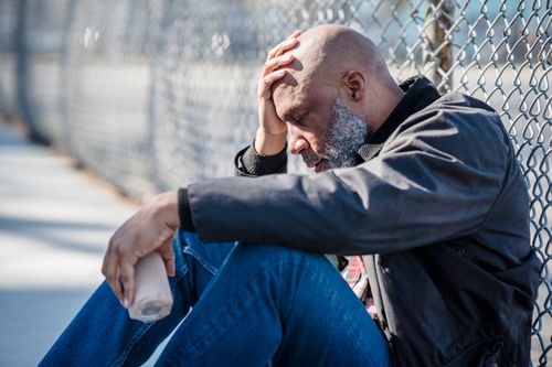Free Pensive adult man with hand on head sitting by a fence, holding bottle. Stock Photo