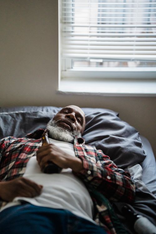 Free Elderly man in casual wear resting on a bed indoors while holding a bottle. Stock Photo