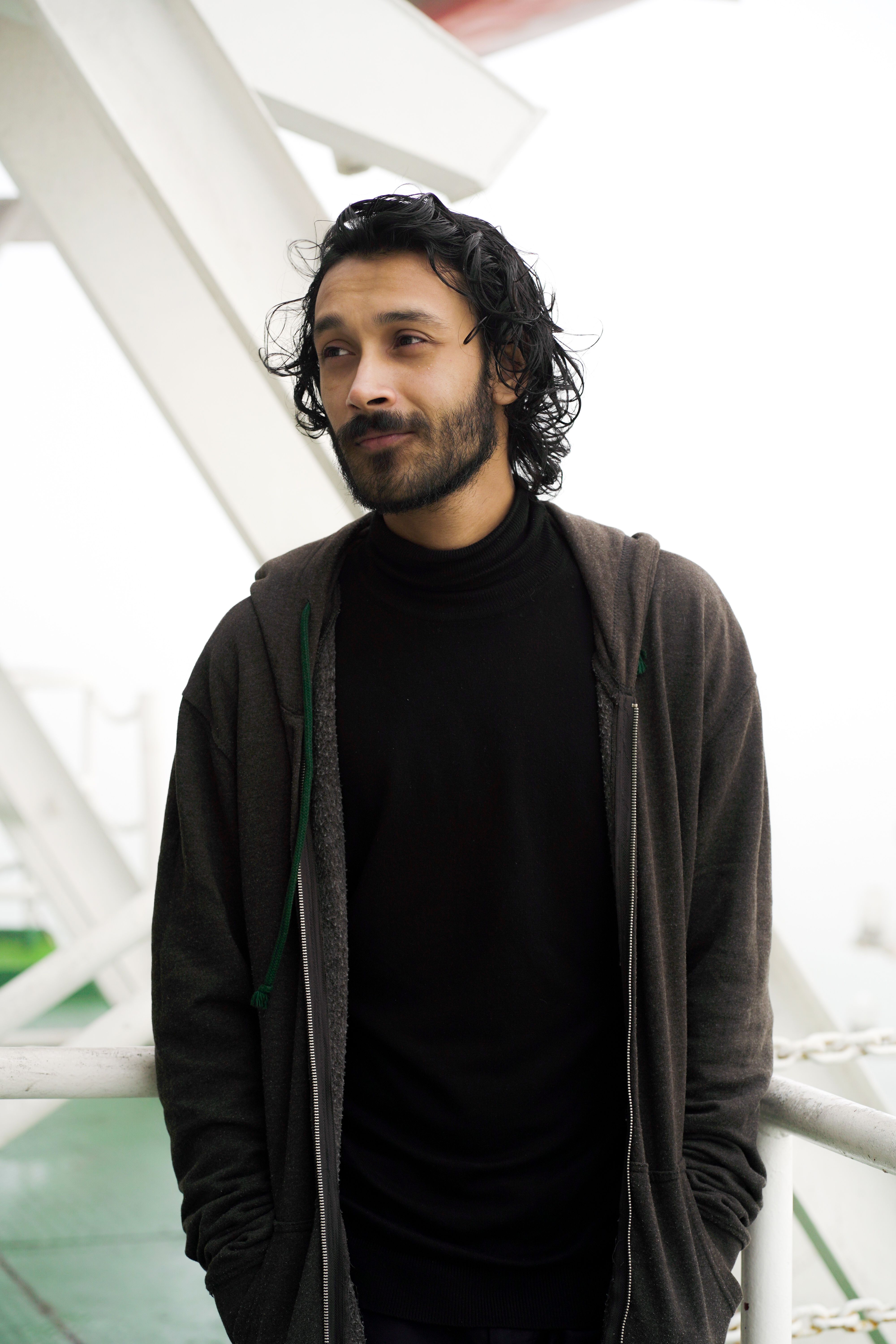 A contemplative man with a beard posing outdoors, hands in pockets.
