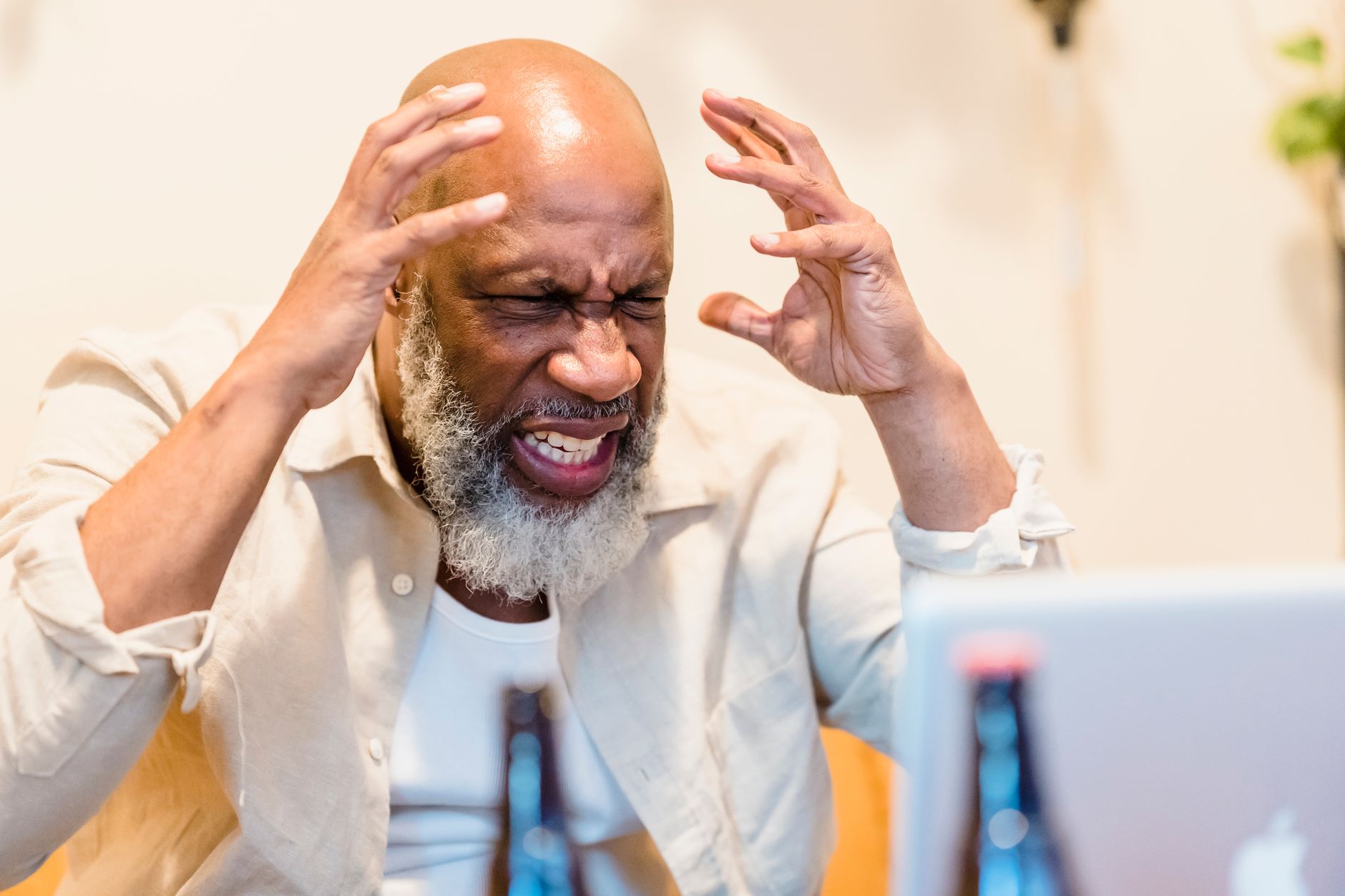 An adult man expressing frustration while sitting in front of a laptop in an indoor setting.