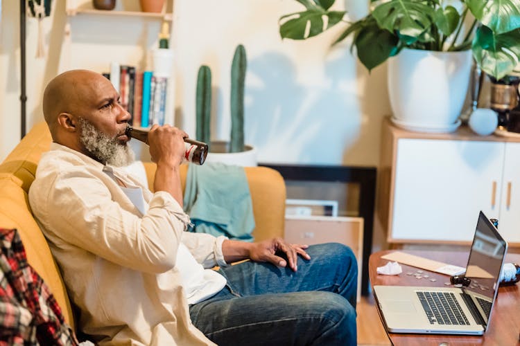 Bald Man With A Gray Beard Sitting Drinking Beer On A Couch In Front Of A Laptop 