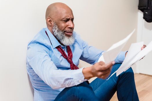 An adult man with a beard looks distressed while sitting on the floor, examining papers.