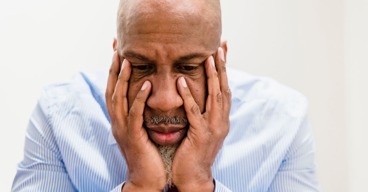 A man with hands to his face against a plain background, looking thoughtful.