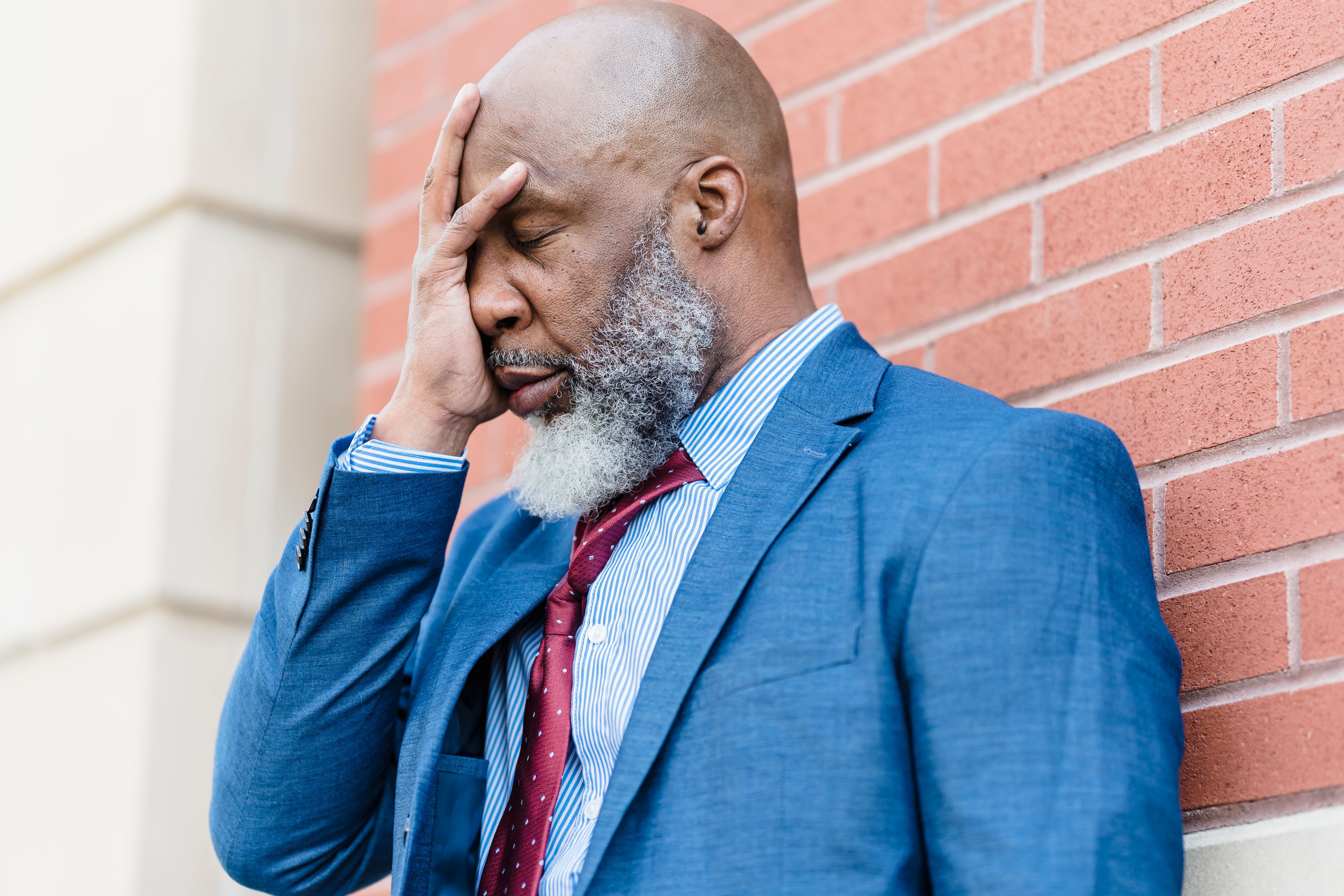 A bearded man in a suit showing emotions while leaning against a wall.