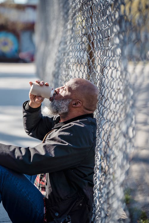 Man Leaning on Chain Link Fence while Drinking