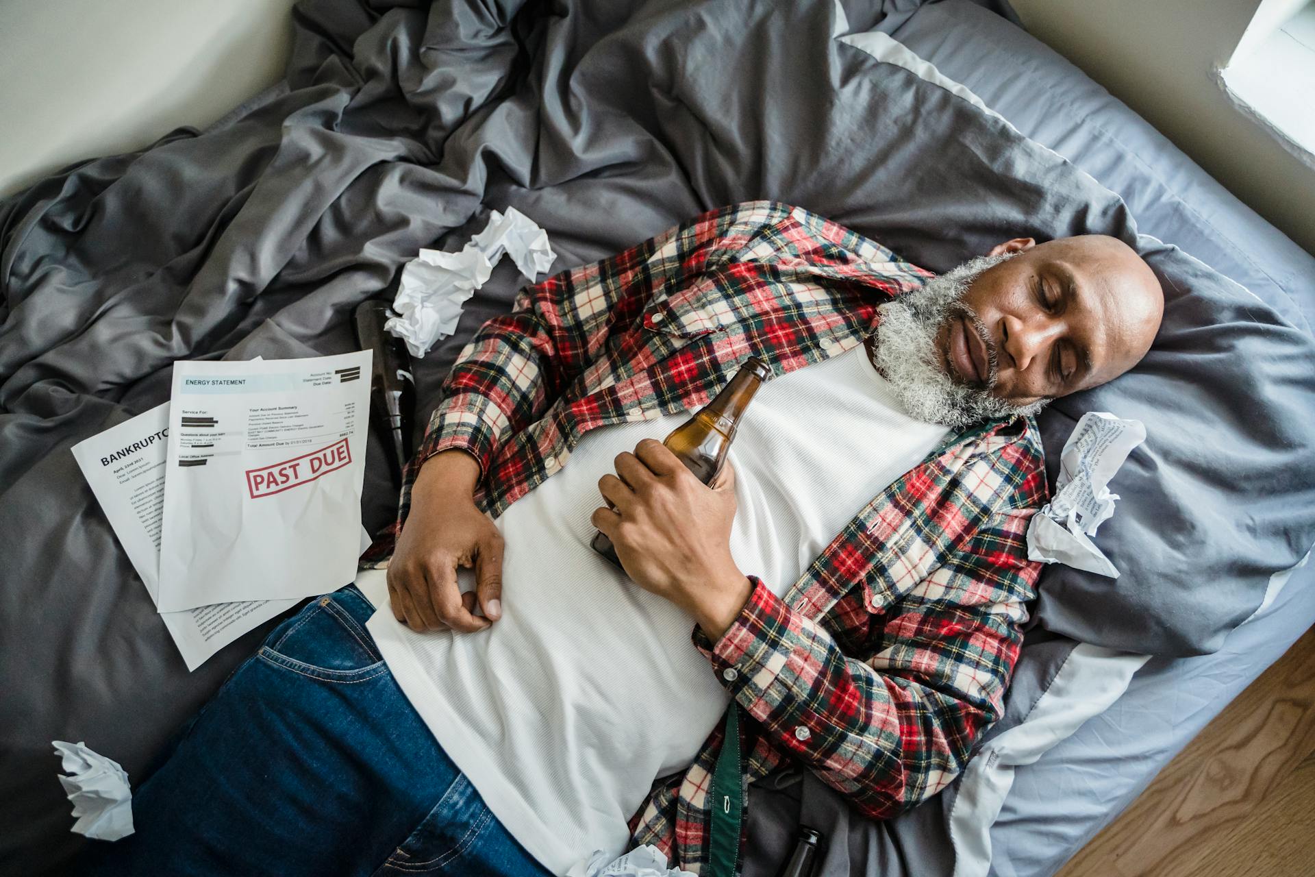 Elderly Man Lying Asleep on a Bed with a Bottle of Beer and Past Due Papers