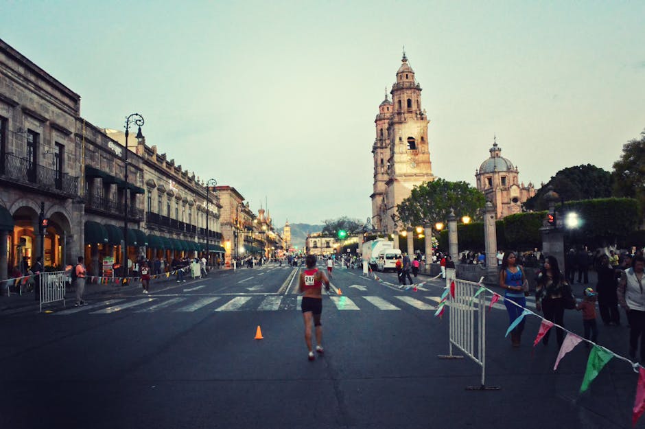 A marathon runner strides through a city street in front of a historic cathedral, captured during daylight amidst a cheering crowd.