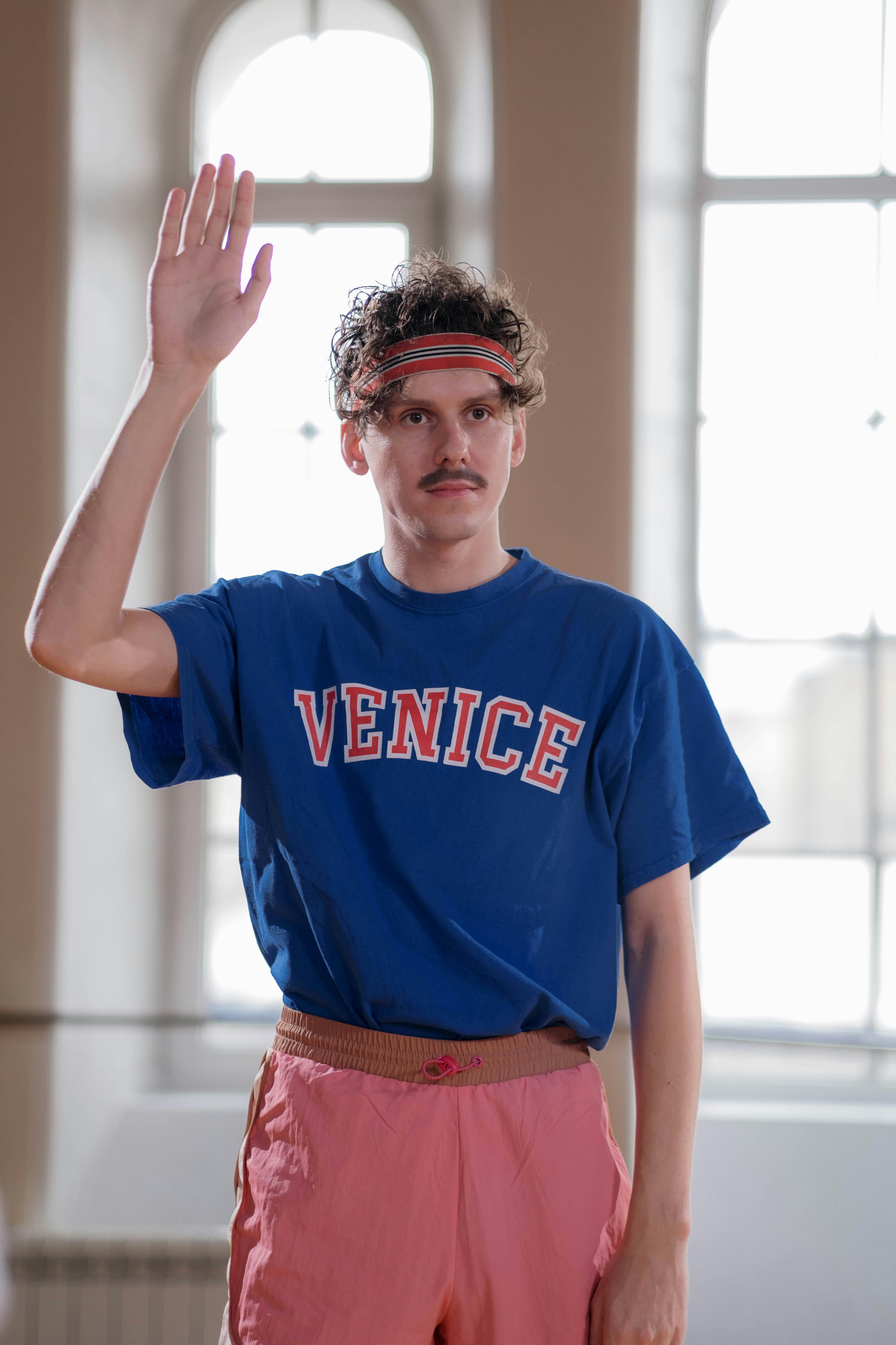 Man with raised arm wearing 'Venice' shirt and headband indoors.